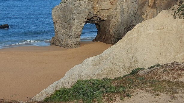 Un paysage côtier caractérisé par une arche rocheuse naturelle, une plage de sable et une vue lointaine sur la mer.