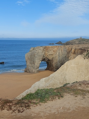 A coastal landscape with a natural rock arch formation, sandy beach, and a distant view of the sea.