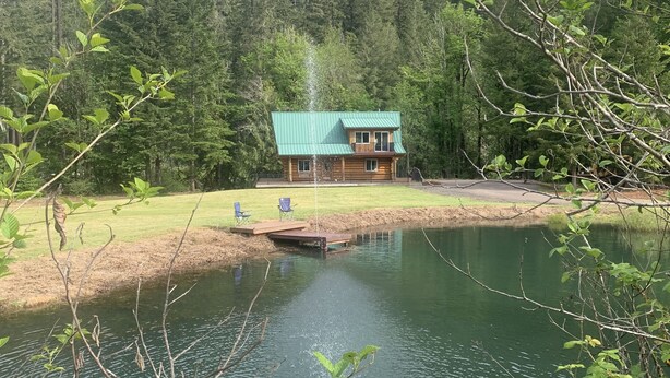 A cabin with a green roof, surrounded by a forest and a lake. - 6 BDRM Log Cabin on Big Fall Creek (Fall Creek)