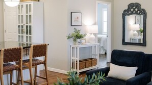 A modern living room with a dark sofa, a dining area with wooden chairs, a white console table with a lamp, and a large mirror on the wall. - Cozy cottage, just a short walk to UofA (Fayetteville)