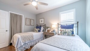A bedroom with two beds, a ceiling fan, a window with blinds, a bedside table with a lamp, and framed artwork on the wall. - Cozy cottage, just a short walk to UofA (Fayetteville)