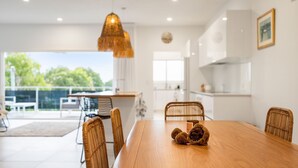 A modern dining area with a wooden table, wicker chairs, and a hanging light fixture. - Sun Crest - amazing ocean views, walk to beach and vibrant village of Woolgoogla (Woolgoolga)