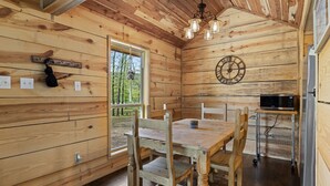 A wooden cabin interior with a dining table, chairs, a kitchen area with a microwave, and a large window providing a view of the outdoors. - Riverside Cabin | Kayaks | Mountains | Stargazing (Nashoba)