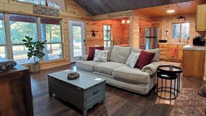 A cozy cabin interior with a sofa, coffee table, and a kitchen area. - Riverside Cabin | Kayaks | Mountains | Stargazing (Nashoba)