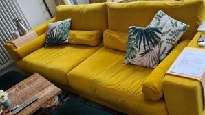 Divano giallo con un portatile, occhiali e un documento, circondato da cuscini a tema tropicale.