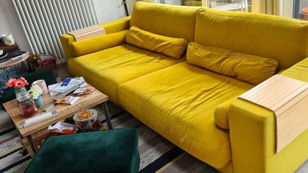 Un divano giallo brillante con cuscini, un tavolino in legno, una poltrona verde e un piccolo tavolo con diversi oggetti.