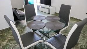 Een eetgedeelte met een glazen tafel en vier stoelen, een bank en een tv op een witte standaard.