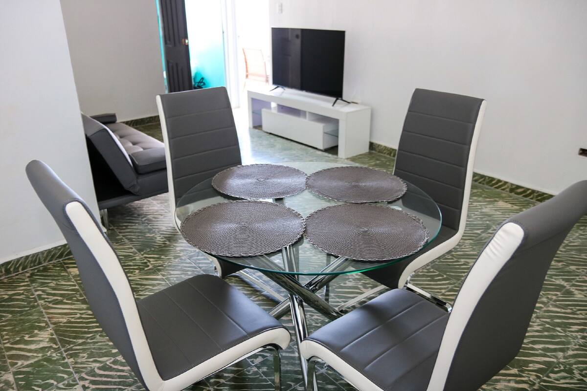 A dining area with a glass table and four chairs, a sofa, and a TV on a white stand.