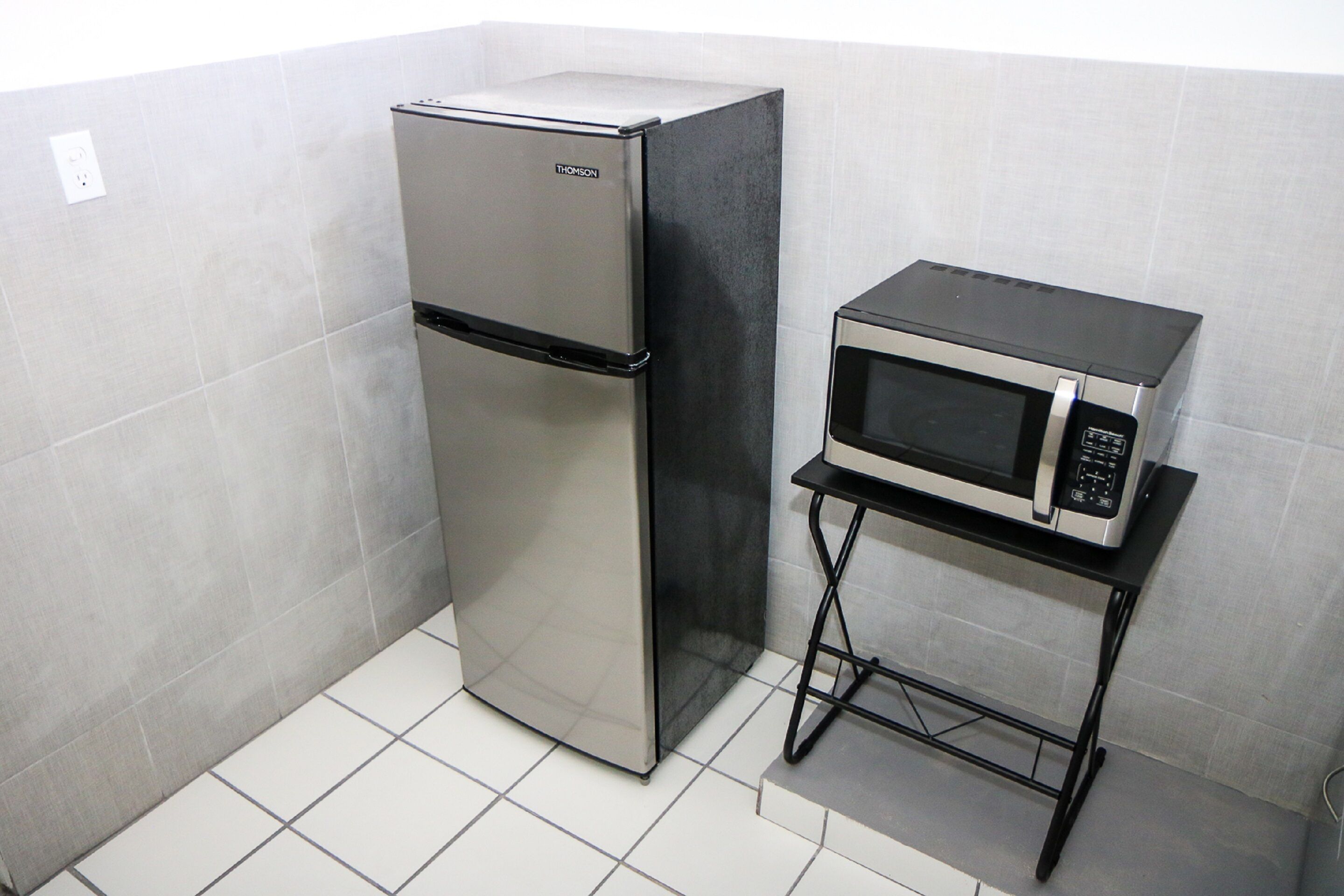 A compact kitchen with a stainless steel refrigerator and a microwave on a small black metal stand.