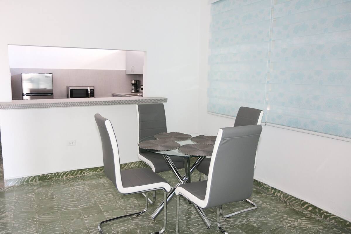 A modern kitchen with stainless steel appliances, a white countertop, and a dining area with a glass table and grey chairs.