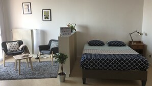 A bedroom with a bed, two chairs, a small table, a potted plant, a bedside lamp, and a sign on the dresser.