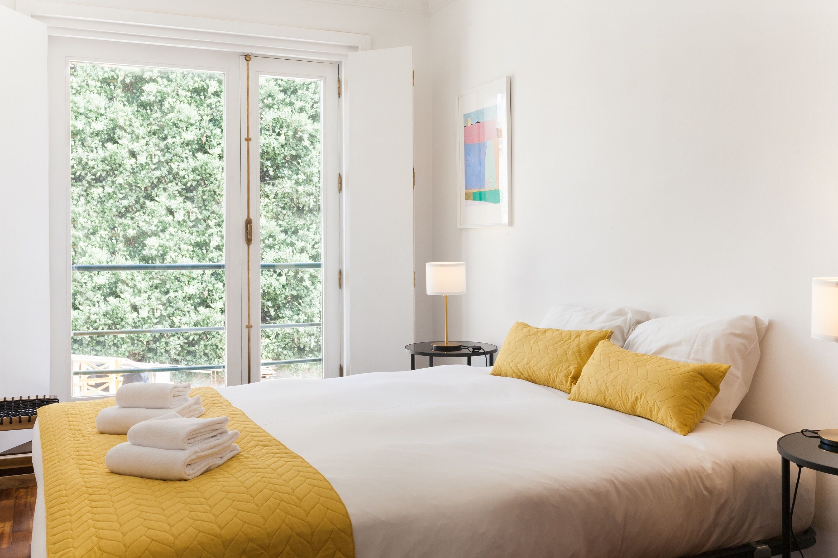 Un cuarto de hotel con una cama, un ventanal con vista a árboles, una mesita de noche con una lámpara y un cuadro en la pared.