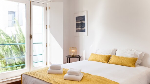 A hotel room with a large bed, a window with a view of greenery, a small table with a lamp, and a framed picture on the wall.