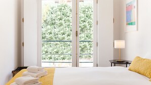 A hotel room with a bed, a window with a view of greenery, a bedside table with a lamp, and a framed picture on the wall.