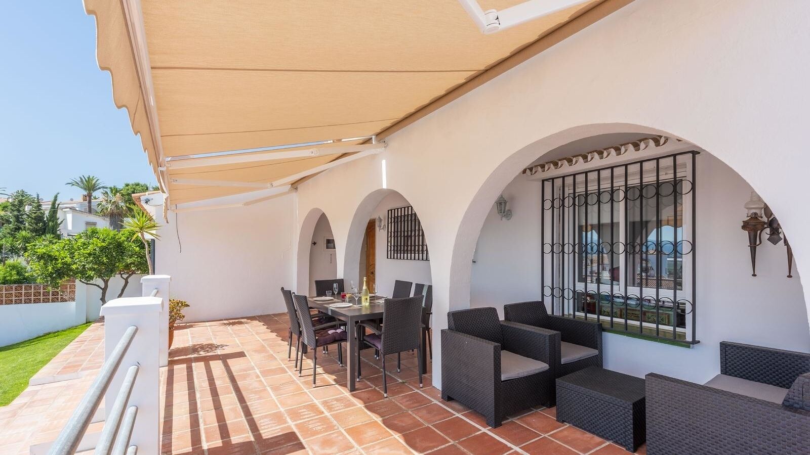 Een overdekt terras met een tafel en stoelen, een bank, en een decoratieve muur met een raam en metalen spijlen.