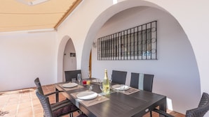 Un comedor con una mesa dispuesta para cuatro personas, rodeada de sillas, bajo una pérgola.