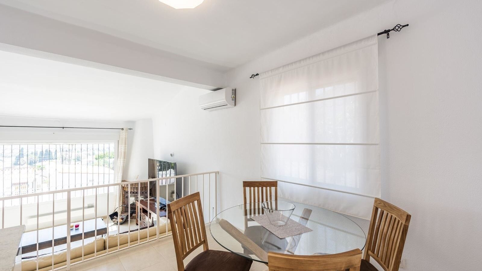 A dining area with a glass table and wooden chairs, a white air conditioning unit on the wall, and a view through a large window.