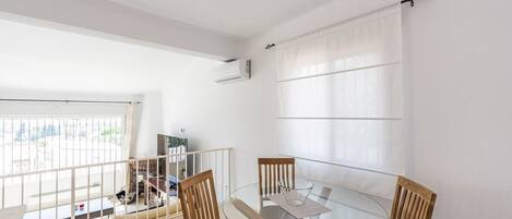A dining area with a glass table and wooden chairs, a white air conditioning unit on the wall, and a view through a large window.