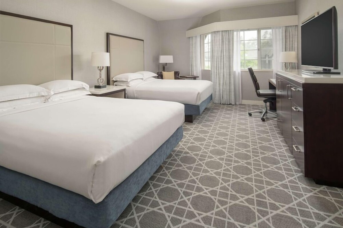 A hotel room with two beds, a desk, a chair, a TV, and a large window with curtains.
