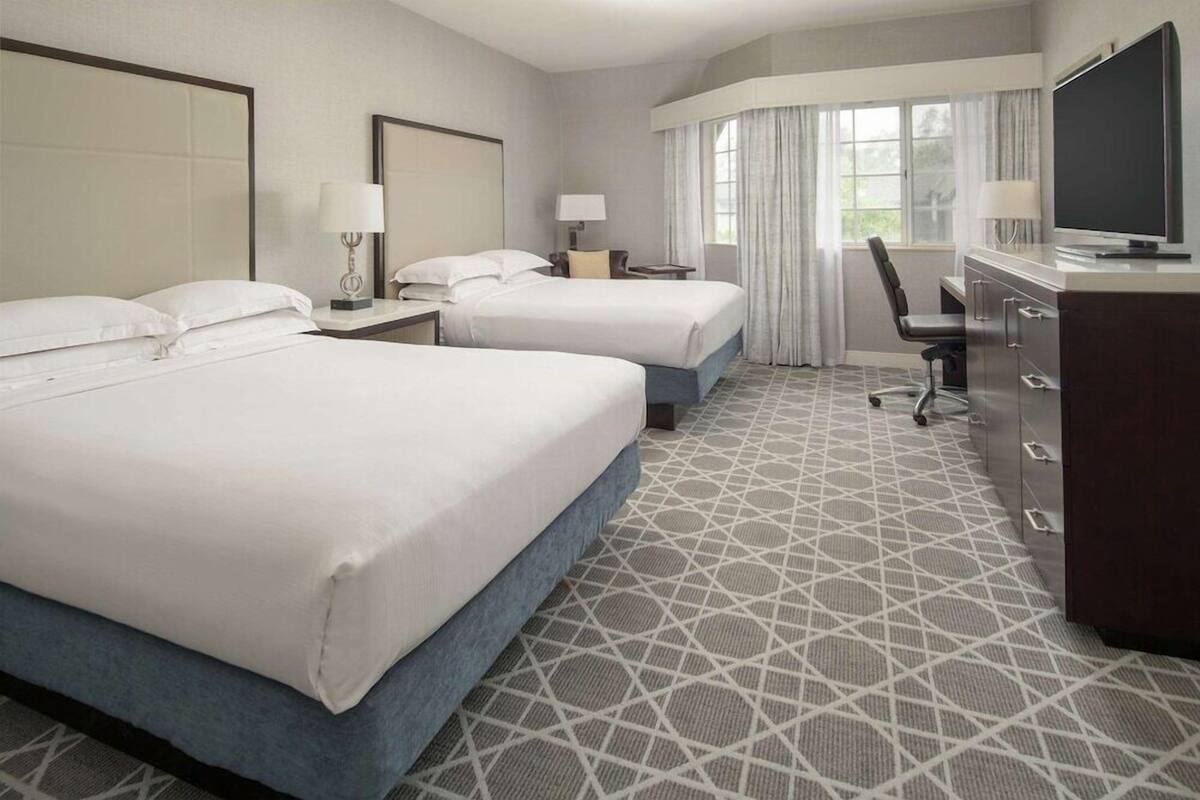 A hotel room with two beds, a desk, a chair, a TV, and a large window with curtains.