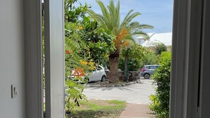 A view through an open door into a garden with a palm tree, a pathway, and parked cars. - Maison 90 m2 Pour 4 Personnes Avec Piscine Collective (Orient baie)