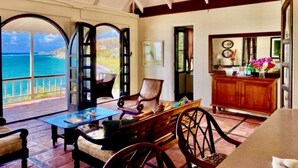 Amplia habitación con un gran ventanal con vista al mar. Hay una mesa de comedor de madera y sillas, una pequeña cocina con un armario de madera, y una zona de estar acogedora con una butaca cómoda.