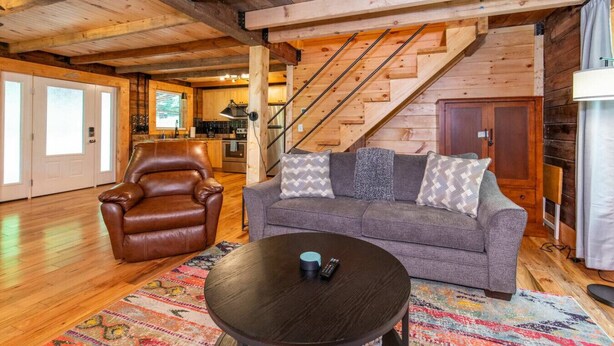 Una sala de estar acogedora con un sillón de cuero, un sofá gris con cojines estampados y una mesita de centro. Una escalera con barandillas de madera conduce a una zona de cocina.