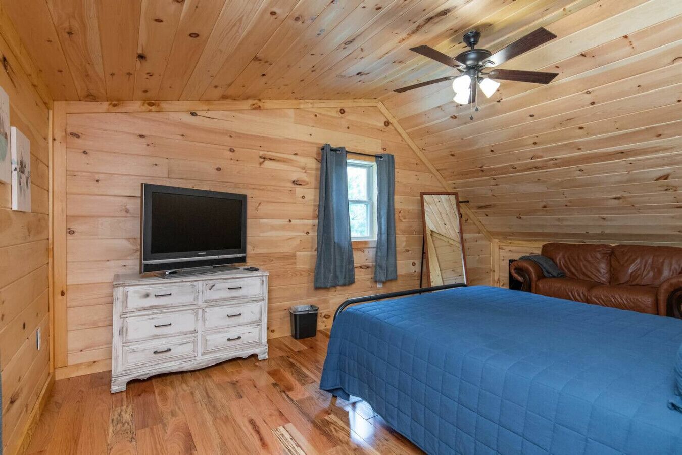 Une chambre avec un plafond en bois, un ventilateur de plafond, un téléviseur posé sur une commode, un lit recouvert d’une couverture bleue, un canapé en cuir marron et une fenêtre avec des rideaux.