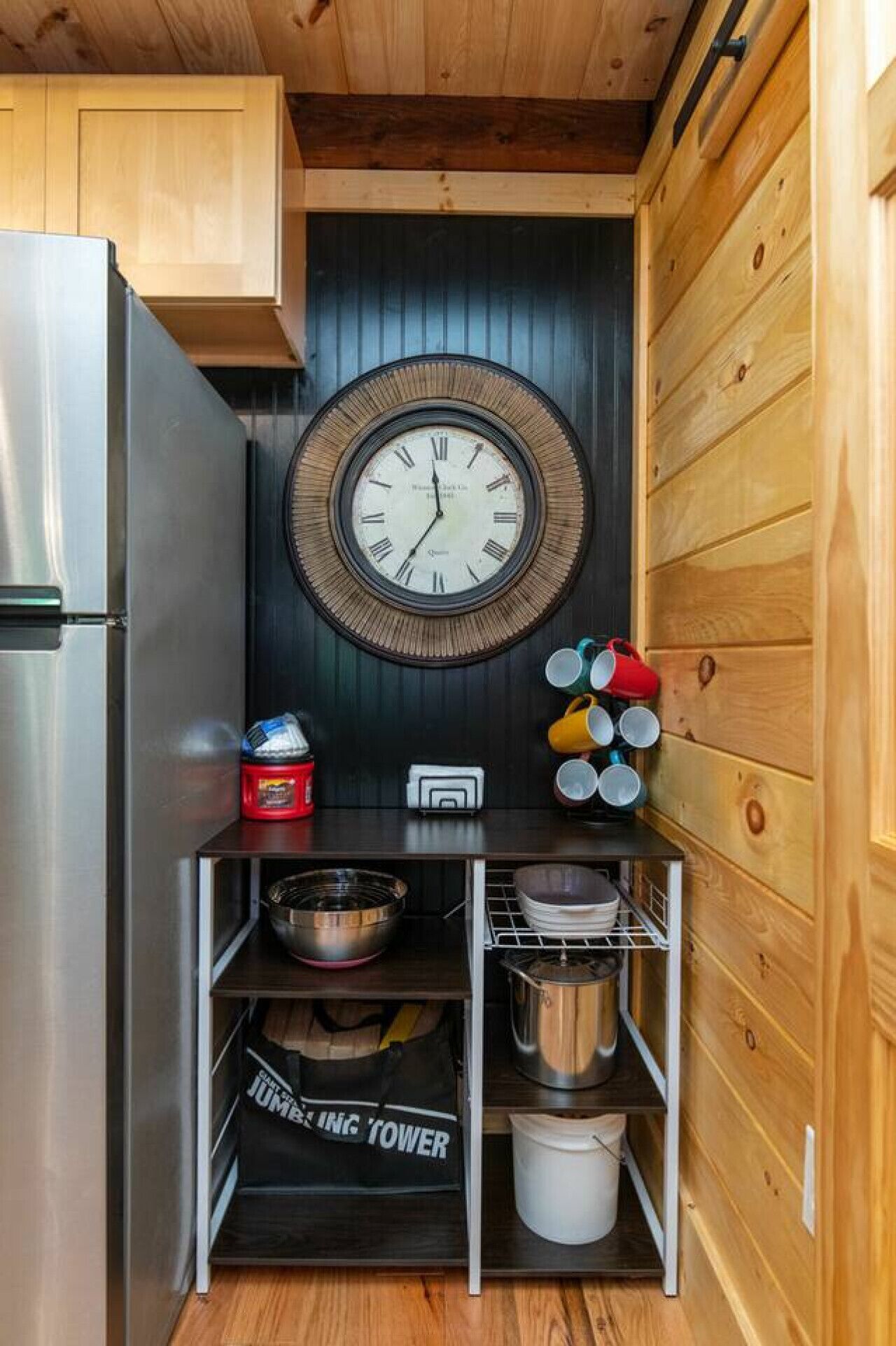 A compact kitchen with a wooden interior, a stainless steel refrigerator, a wall clock, and a shelf with kitchen utensils and a bag.