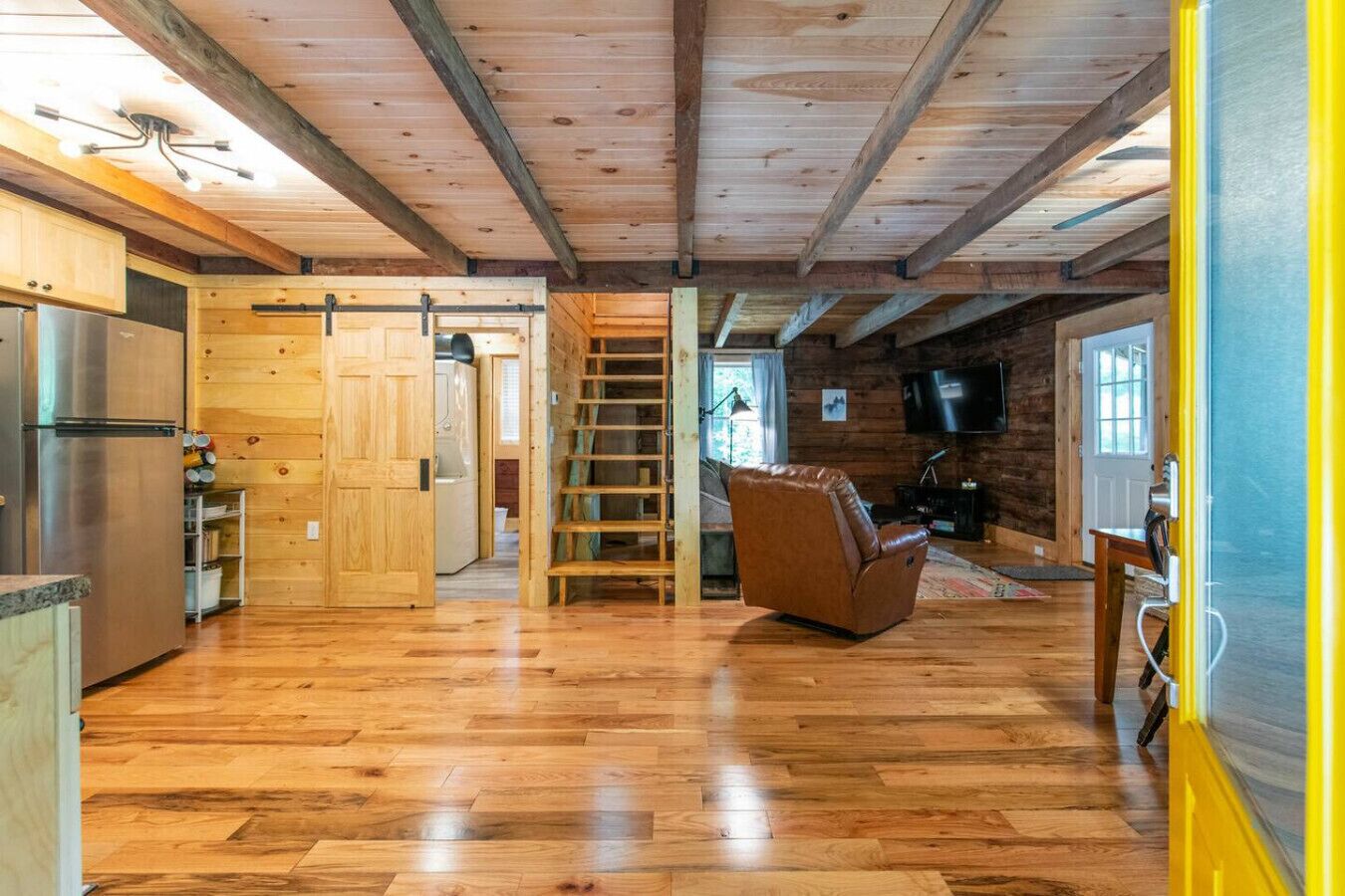 Una habitación amplia con pisos de madera, un ventanal grande, televisión y una escalera que lleva a un altillo.