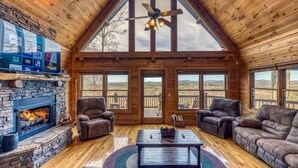 A log cabin interior with a stone fireplace, leather sofas, a ceiling fan, and large windows offering views of the outdoors. - In The Heights - Hot Tub | Fire Pit | Game Room | Mountain Views (Morganton)