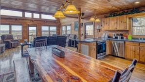 A spacious living area with a wooden dining table, leather sofas, and a large window offering a view of the mountains. - In The Heights - Hot Tub | Fire Pit | Game Room | Mountain Views (Morganton)
