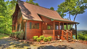 A wooden cabin with a porch, surrounded by trees and a clear sky. - In The Heights - Hot Tub | Fire Pit | Game Room | Mountain Views (Morganton)