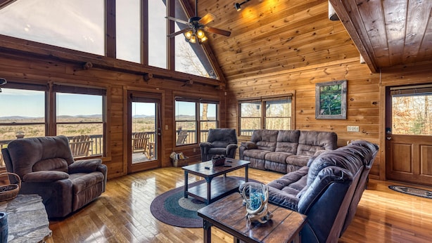 A spacious living room with wooden walls and large windows, furnished with leather sofas and armchairs, and a central coffee table. - In The Heights - Hot Tub | Fire Pit | Game Room | Mountain Views (Morganton)