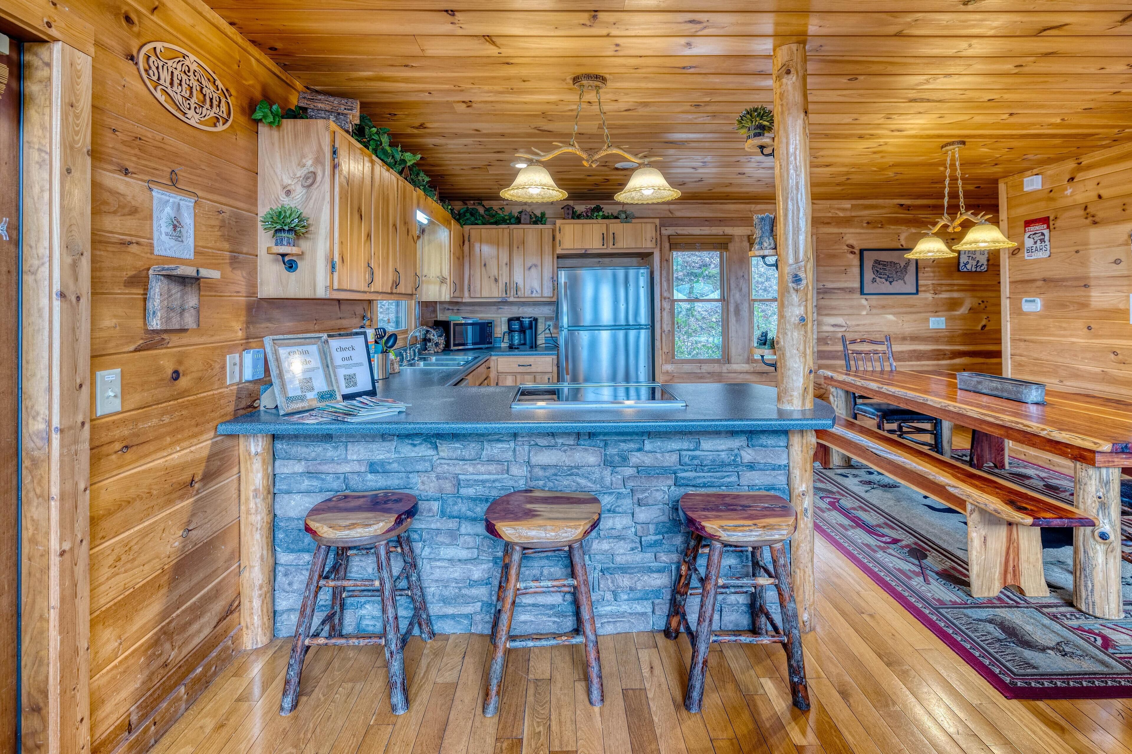 A rustic cabin interior with wooden walls, a stone countertop, a stainless steel refrigerator, and a bar area with stools.