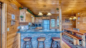 A rustic cabin interior with wooden walls, a stone countertop, a stainless steel refrigerator, and a bar area with stools. - In The Heights - Hot Tub | Fire Pit | Game Room | Mountain Views (Morganton)