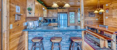 A rustic cabin interior with wooden walls, a stone countertop, a stainless steel refrigerator, and a bar area with stools.