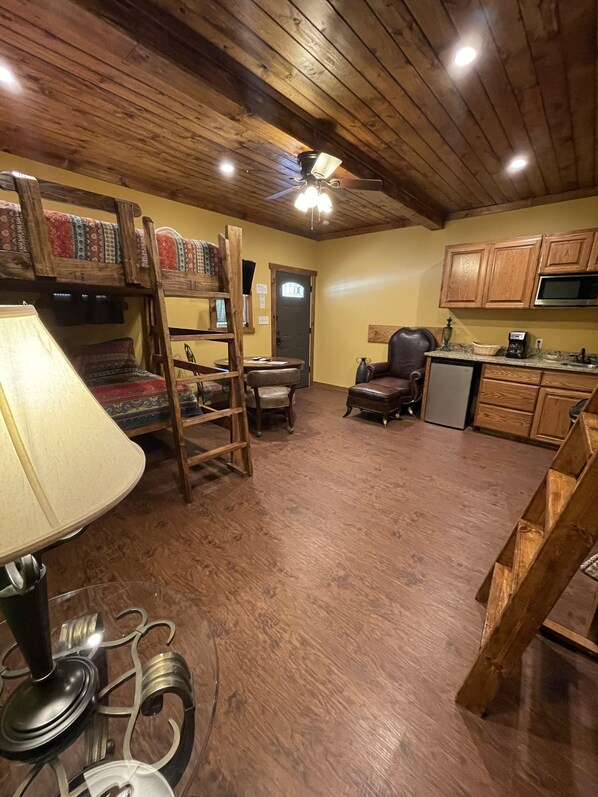 Una habitación acogedora con pisos de madera, un ventilador de techo, una mesa de comedor con sillas, un sofá y una zona de cocina con armarios de madera y un microondas.