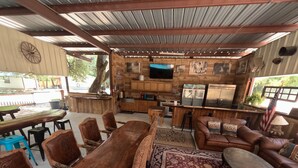 A rustic cabin interior with a long wooden dining table, leather sofas, and a kitchen area. - Haggville Hacienda - Private Riverfront @Vacation Frio River - Pool Sleeps 29-38 (Concan)
