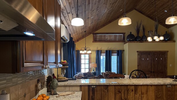 A kitchen with a wooden ceiling, granite countertop, and a large island. There are fruits on the counter and a hanging light fixture. - Haggville Hacienda - Private Riverfront @Vacation Frio River - Pool Sleeps 29-38 (Concan)