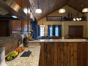 Una cocina con techo de madera, encimera de granito y una isla grande. Hay frutas sobre la encimera y una lámpara colgante.