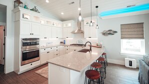 Een moderne keuken met witte kastjes, roestvrijstalen apparatuur, een centraal eiland met krukken, hanglampen en een raam met uitzicht.