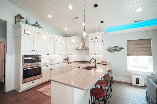 Una cocina moderna con armarios blancos, electrodomésticos de acero inoxidable, una isla central con taburetes, iluminación colgante y un ventanal con vistas.
