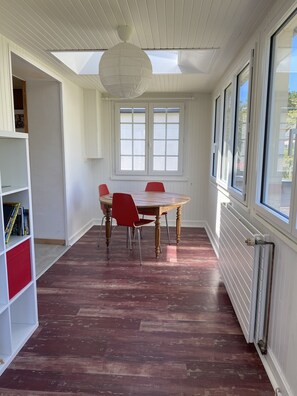 A dining area with a wooden table and red chairs, large windows, and a white ceiling.
