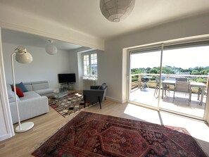 Un salon spacieux avec une grande fenêtre, un canapé gris, un téléviseur à écran plat et un tapis à motifs.