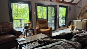 A spacious living room with a brown leather sofa, a wooden coffee table, and a large window with a view of trees. - Rustic, Cozy Cabin with a View of the Smokies, Firepit, Large Deck, Pet-Friendly (Almond)