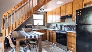 A kitchen with wooden cabinets, a black refrigerator, a microwave, a stove, and a dining area with a table and chairs. - Rustic, Cozy Cabin with a View of the Smokies, Firepit, Large Deck, Pet-Friendly (Almond)