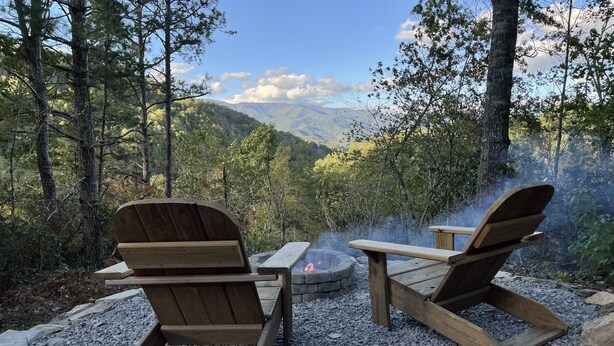 Two Adirondack chairs facing a campfire in a forested area with a view of mountains. - Rustic, Cozy Cabin with a View of the Smokies, Firepit, Large Deck, Pet-Friendly (Almond)