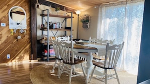 Uma sala de jantar com mesa de madeira e cadeiras brancas, um tapete tecido e uma estante com diversos objetos.