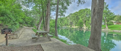 Zona de picnic con bancos y un sendero de hormigón que lleva a un lago tranquilo, rodeado de árboles altos.